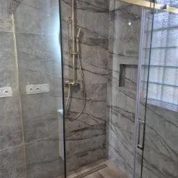 Nowoczesna kabina prysznicowa z przesuwnymi drzwiami, wykończona płytkami imitującymi marmur w odcieniach szarości, z złotą armaturą i odpływem liniowym.