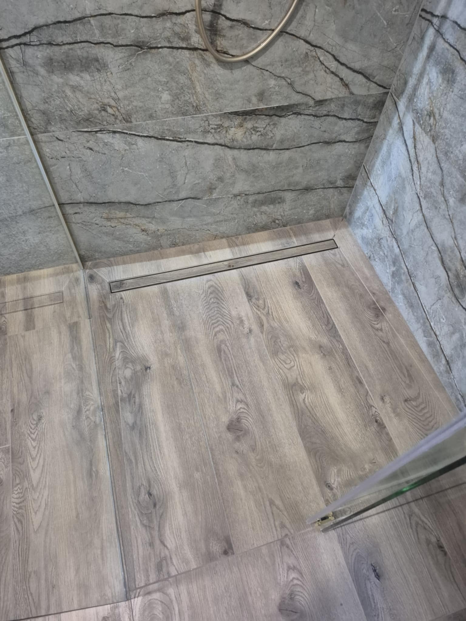 Nowoczesna kabina prysznicowa z odpływem liniowym, wyłożona płytkami imitującymi drewno na podłodze i szarymi płytkami z wzorem marmuru na ścianach.