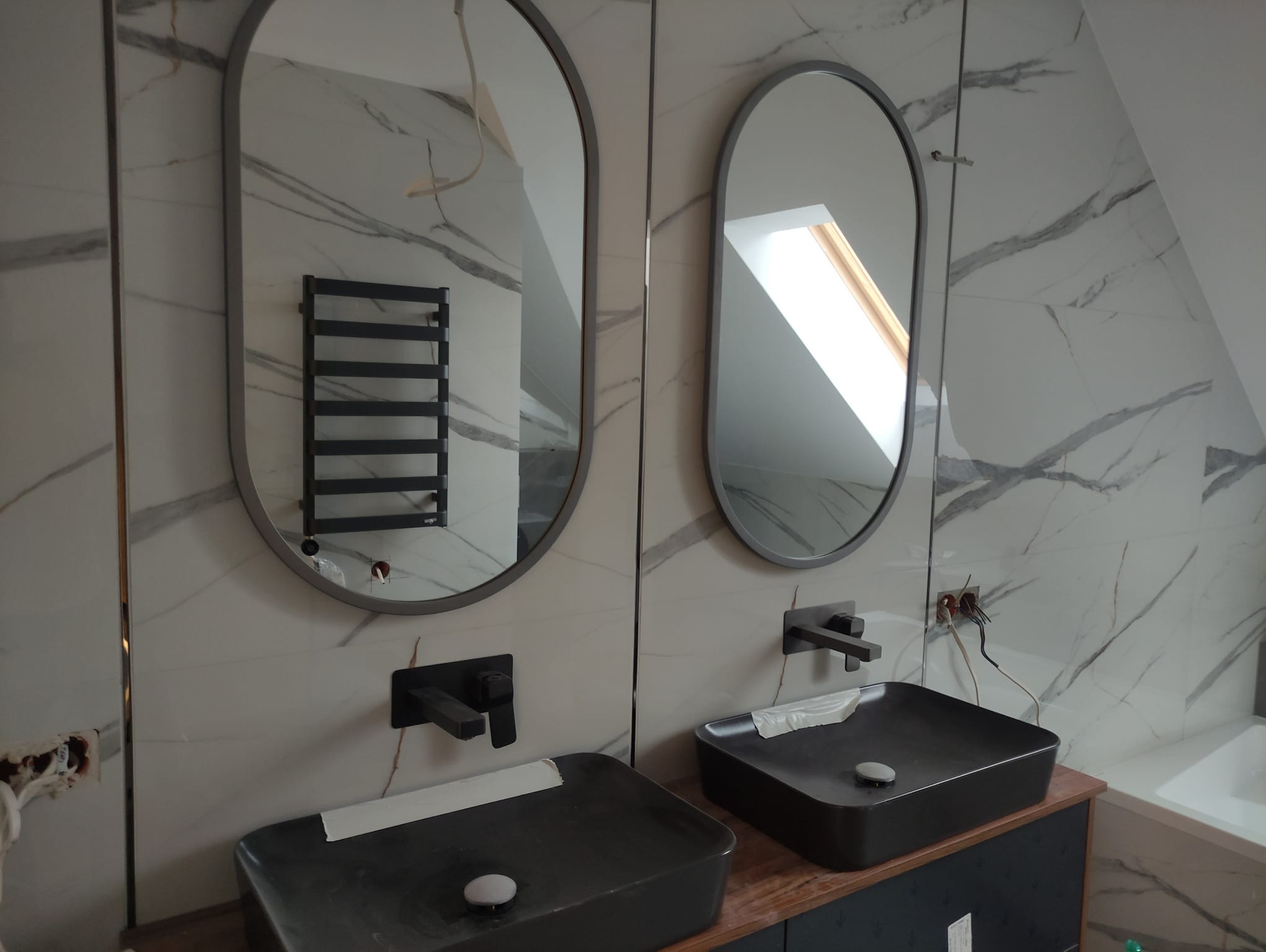 Nowoczesna łazienka w trakcie wykańczania: dwie czarne umywalki nablatowe z czarnymi bateriami, owalne lustra w szarych ramach, białe płytki imitujące marmur, widoczne przewody elektryczne.
