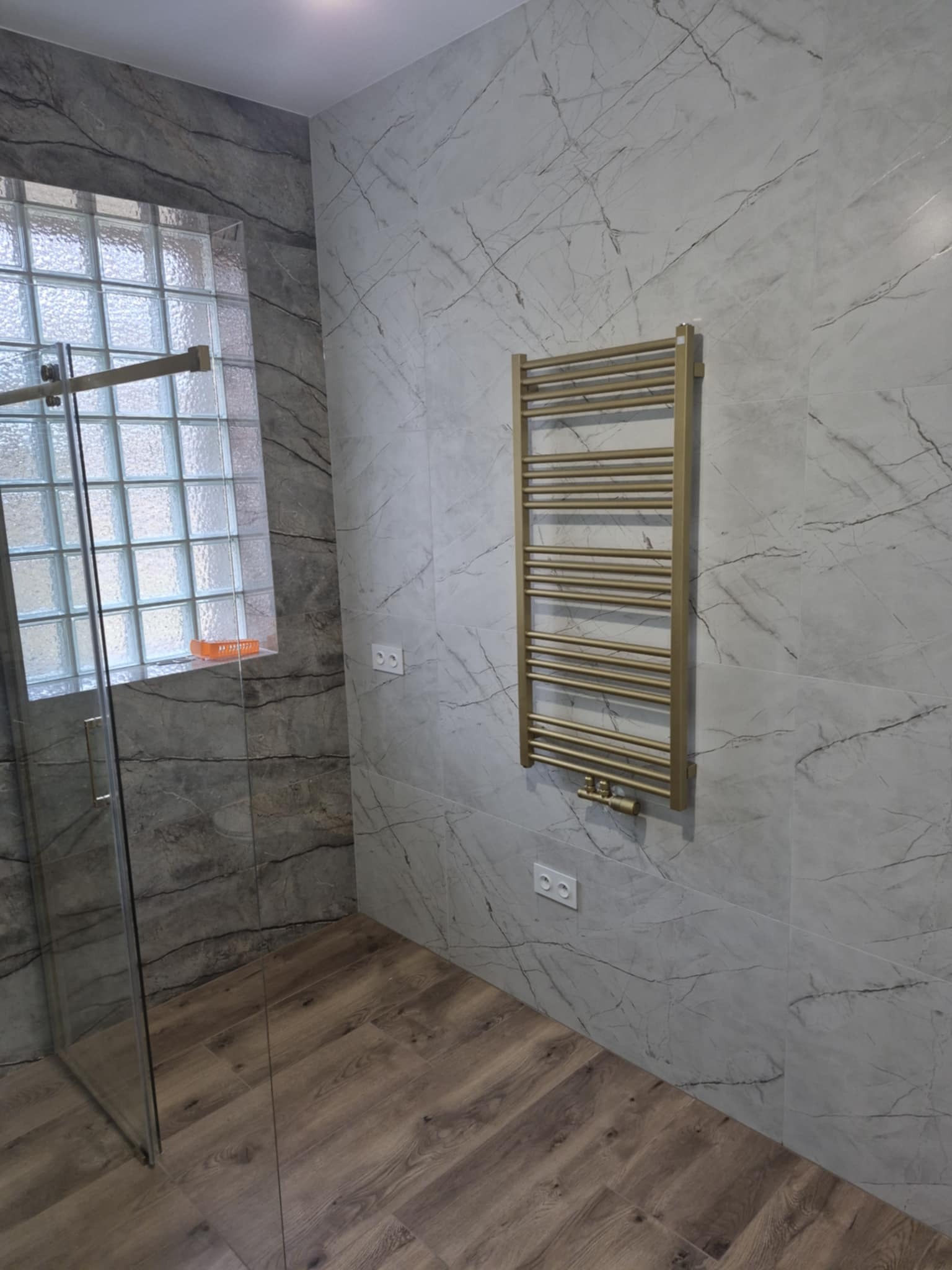 Nowoczesna łazienka z szarymi płytkami imitującymi marmur, drewnopodobną podłogą, złotym grzejnikiem drabinkowym i kabiną prysznicową z luksferami w tle.