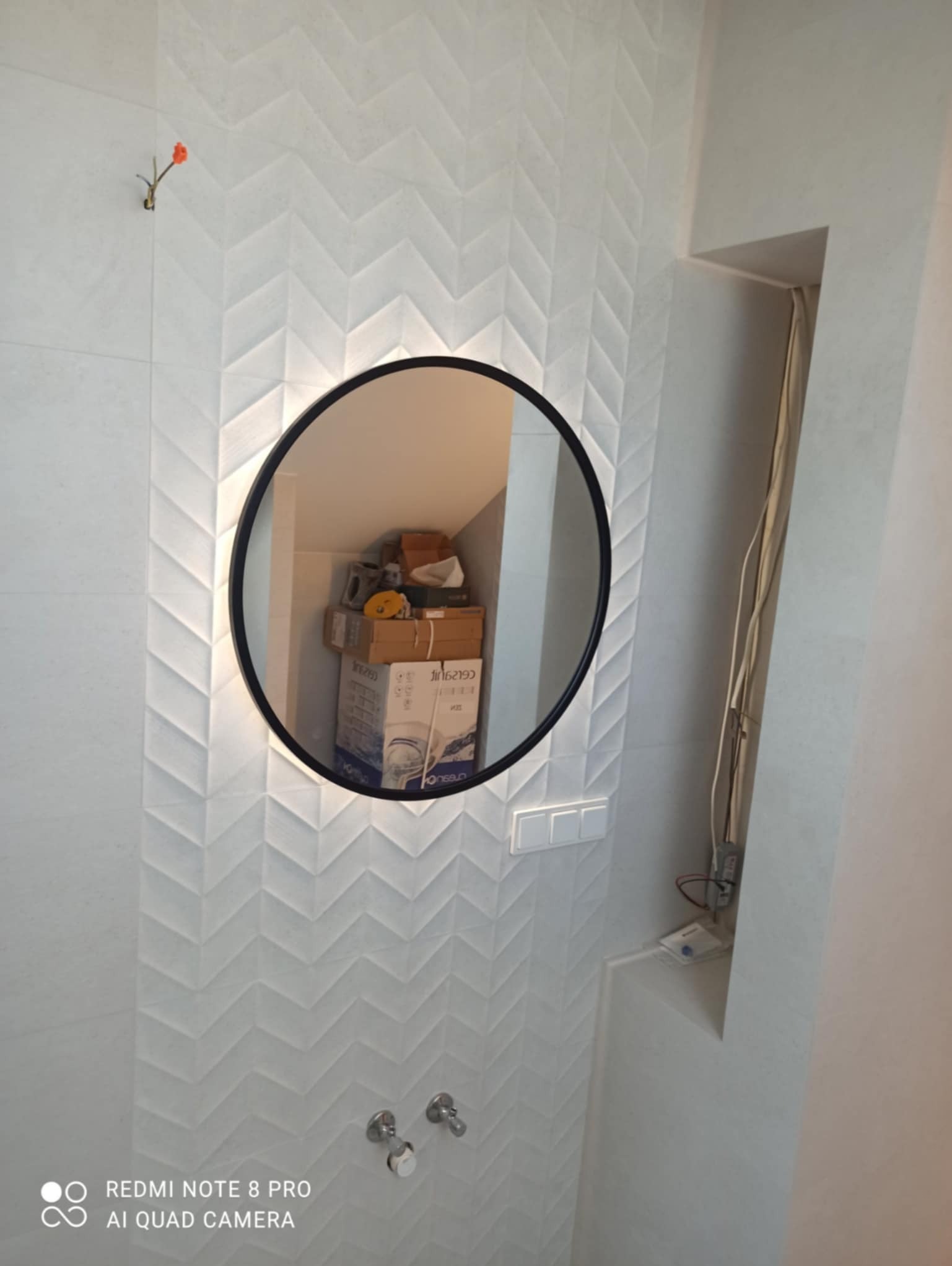 Biała łazienka z wzorzystymi płytkami, okrągłe lustro z podświetleniem odbija kartony na poddaszu, widoczne dwa krany i przełącznik światła.