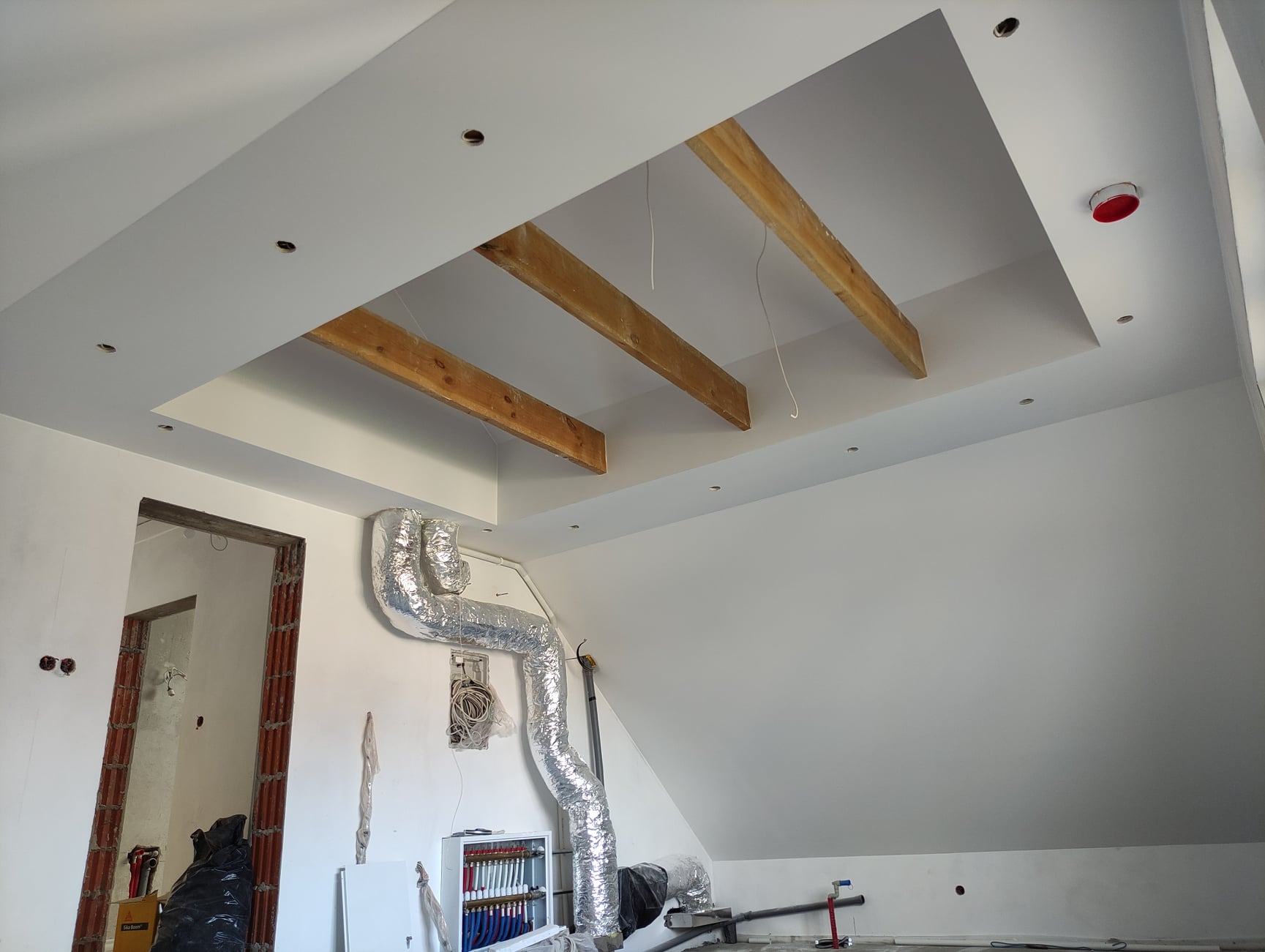 Wnętrze poddasza w trakcie prac wykończeniowych: widoczne drewniane belki w suficie, instalacja wentylacyjna w srebrnej folii, ściany przygotowane pod malowanie, otwór drzwiowy w stanie surowym.
