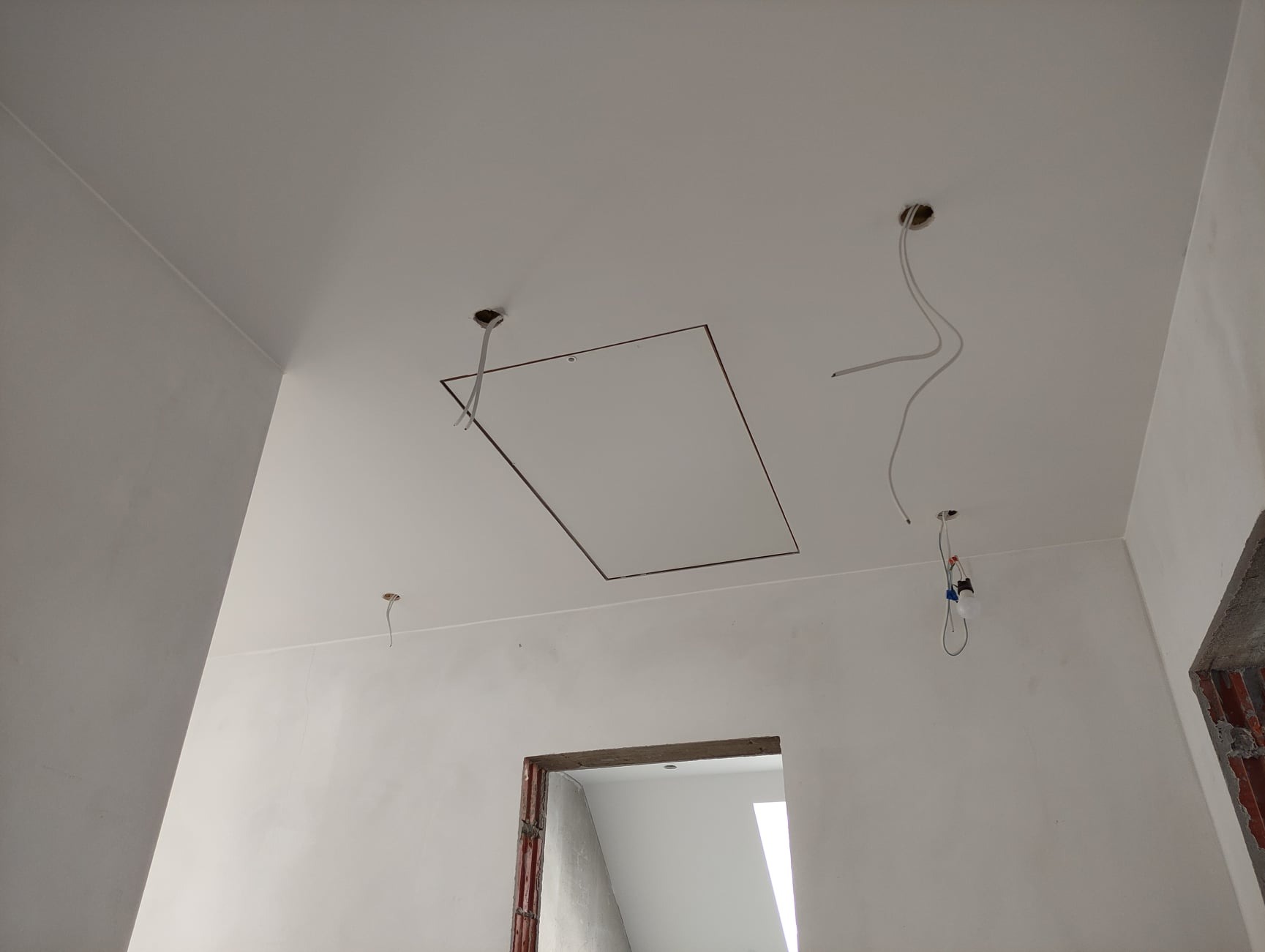 Białe ściany i sufit w trakcie prac wykończeniowych, widoczne otwory na oświetlenie i rama otworu rewizyjnego w suficie, zwisające kable elektryczne, surowy otwór drzwiowy w tle.