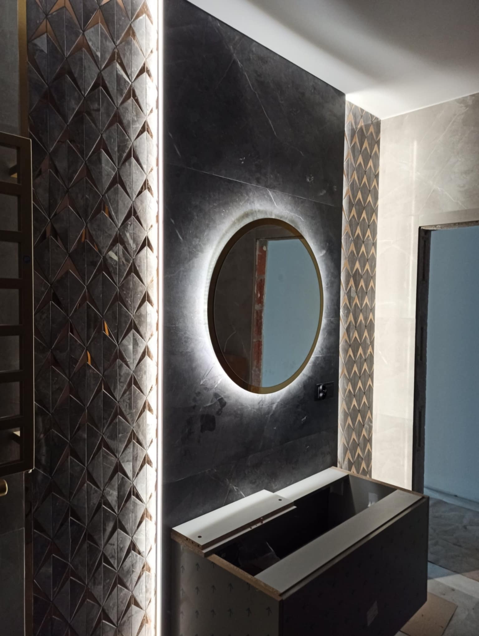 Łazienka w trakcie remontu: ściana z czarnych płytek imitujących marmur, dekoracyjny panel z mozaiką w geometryczne wzory, okrągłe lustro z podświetleniem LED, niedokończona szafka pod umywalkę.