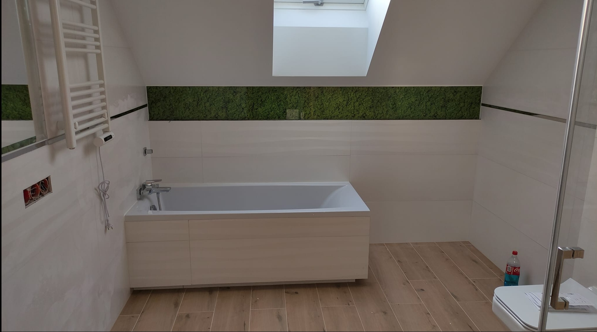 Nowoczesna łazienka na poddaszu z białą wanną obudowaną drewnem, płytkami imitującymi drewno na podłodze i paskiem zielonego mchu na ścianie pod oknem dachowym.