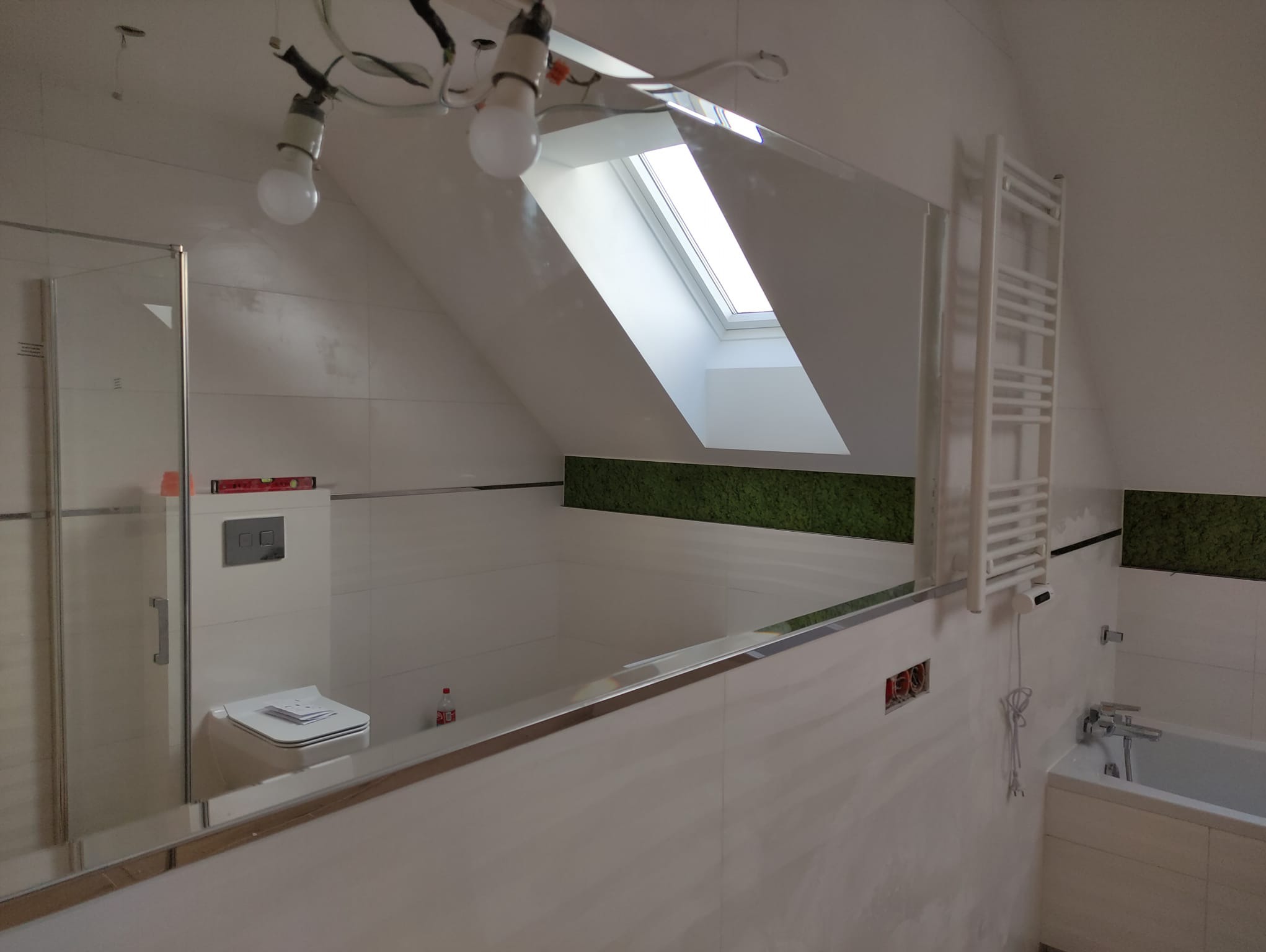 Łazienka w trakcie remontu: widoczna biała glazura, wanna, toaleta, grzejnik drabinkowy, okno dachowe i niezabezpieczone kable oświetleniowe.