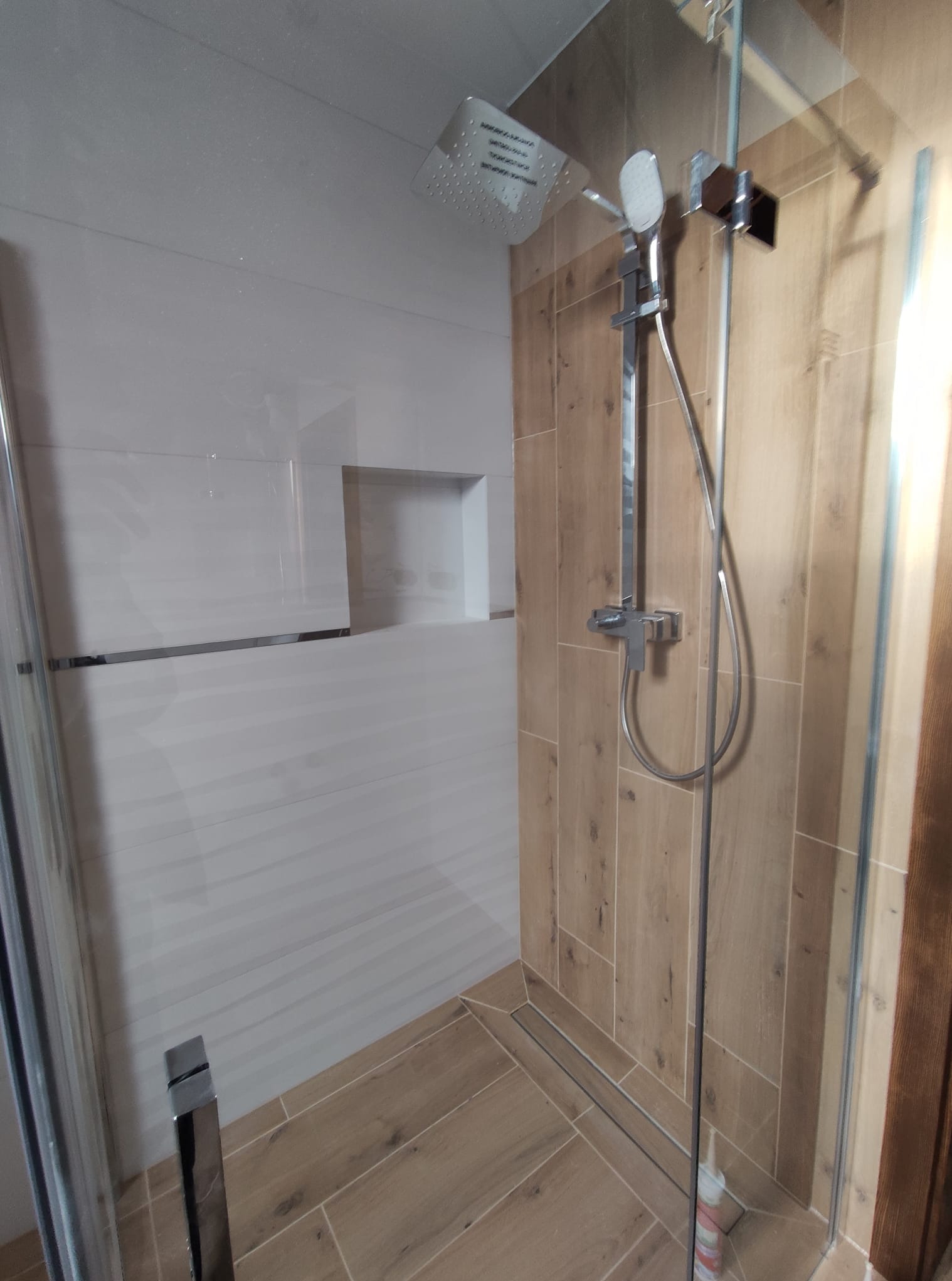 Nowoczesna kabina prysznicowa z deszczownicą i panelami ściennymi imitującymi drewno, widok z boku, minimalistyczny design z wnęką w ścianie wyłożonej białymi płytkami.