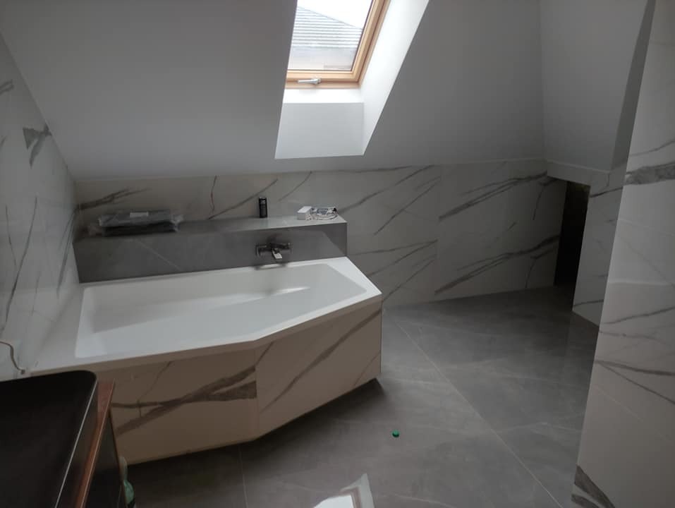 Jasna łazienka na poddaszu z oknem dachowym, wykończona płytkami imitującymi marmur z szarymi żyłami, z narożną wanną obudowaną tymi samymi płytkami i szarą półką nad wanną.