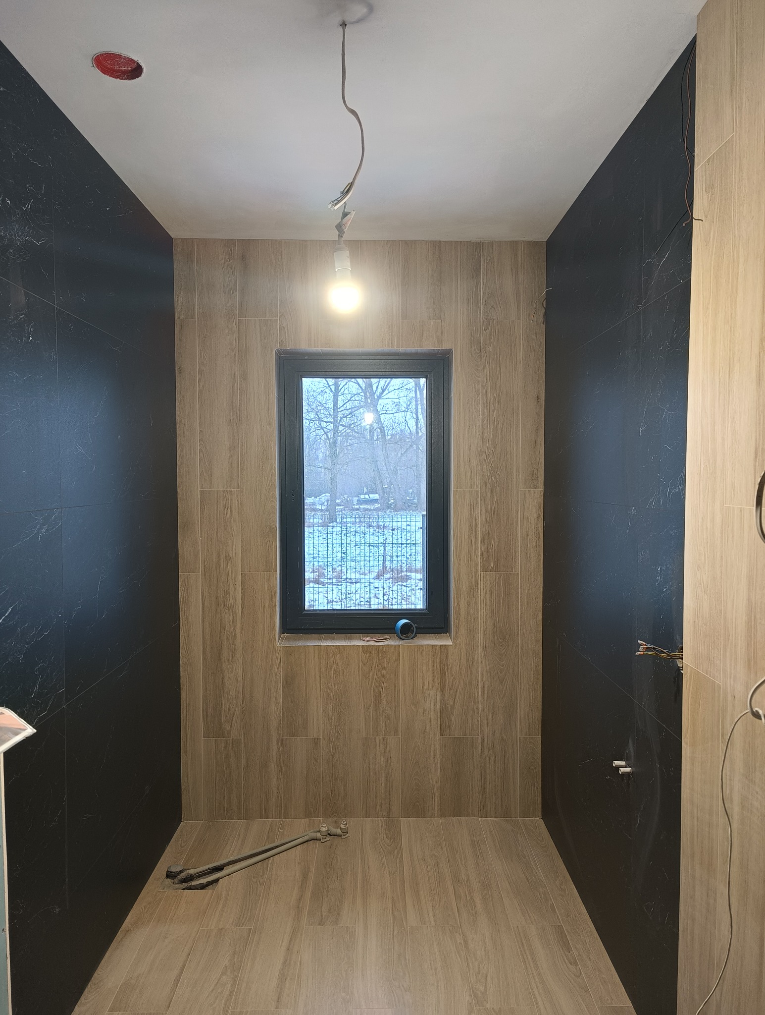 Wnętrze łazienki w trakcie remontu z czarnymi i drewnopodobnymi płytkami, widoczne narzędzia na podłodze i okno z widokiem na zimowy krajobraz.