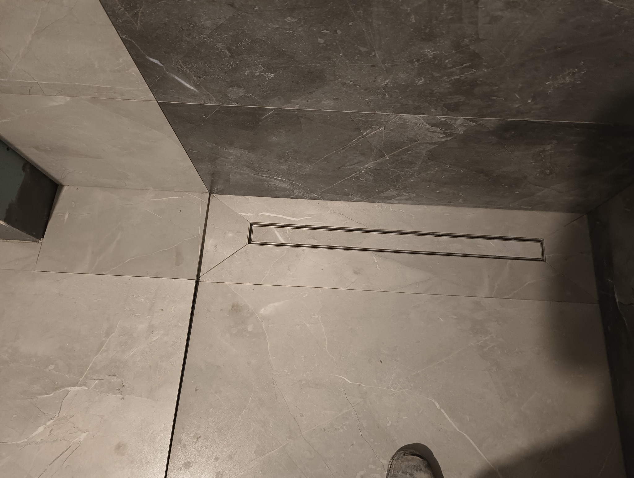 Szare płytki podłogowe i ścienne w łazience z odpływem liniowym, widoczny fragment buta.