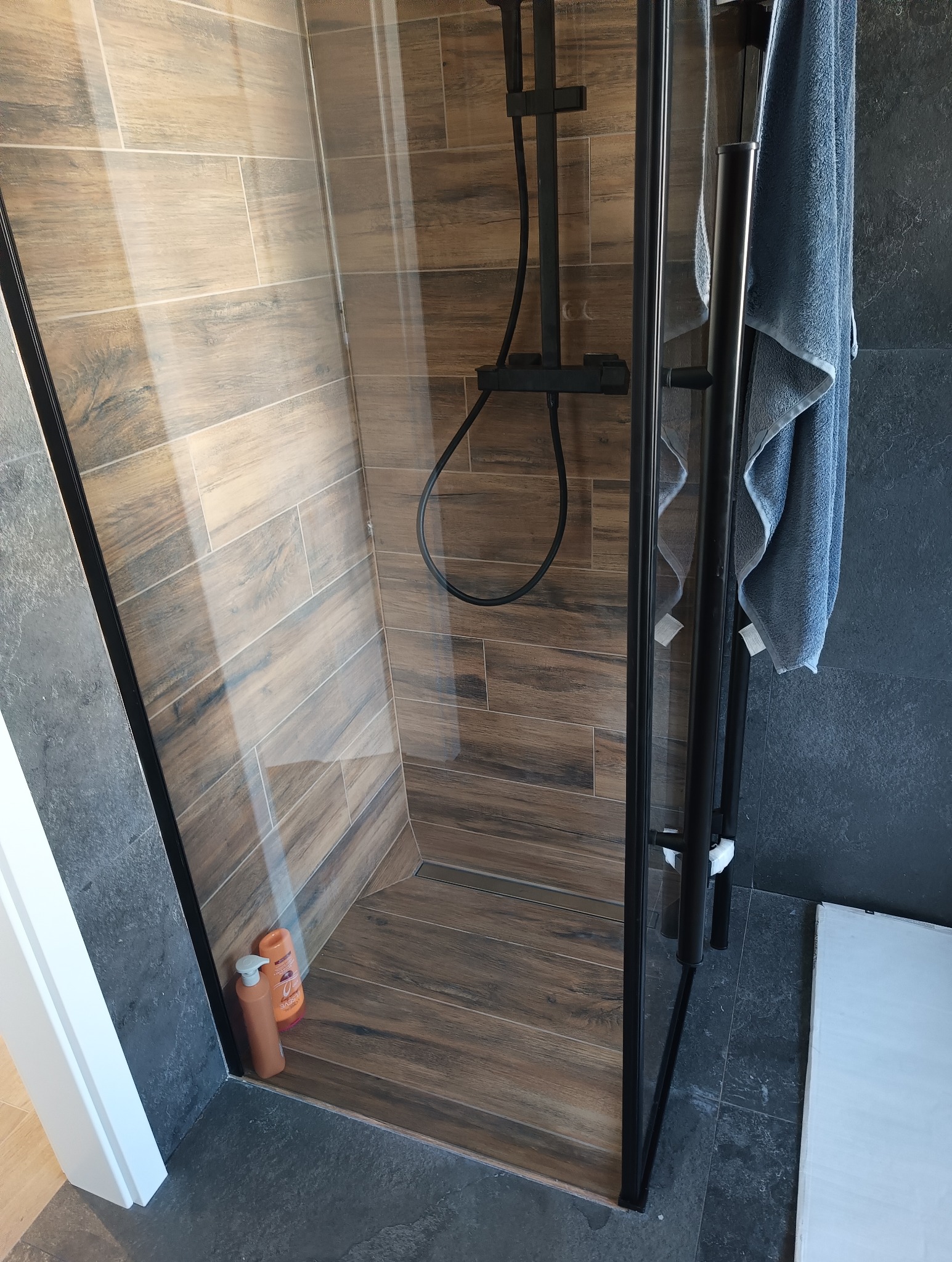 Kabina prysznicowa z drewnopodobnymi płytkami, czarną armaturą i szklanymi drzwiami, widok z góry, butelki kosmetyków w rogu.