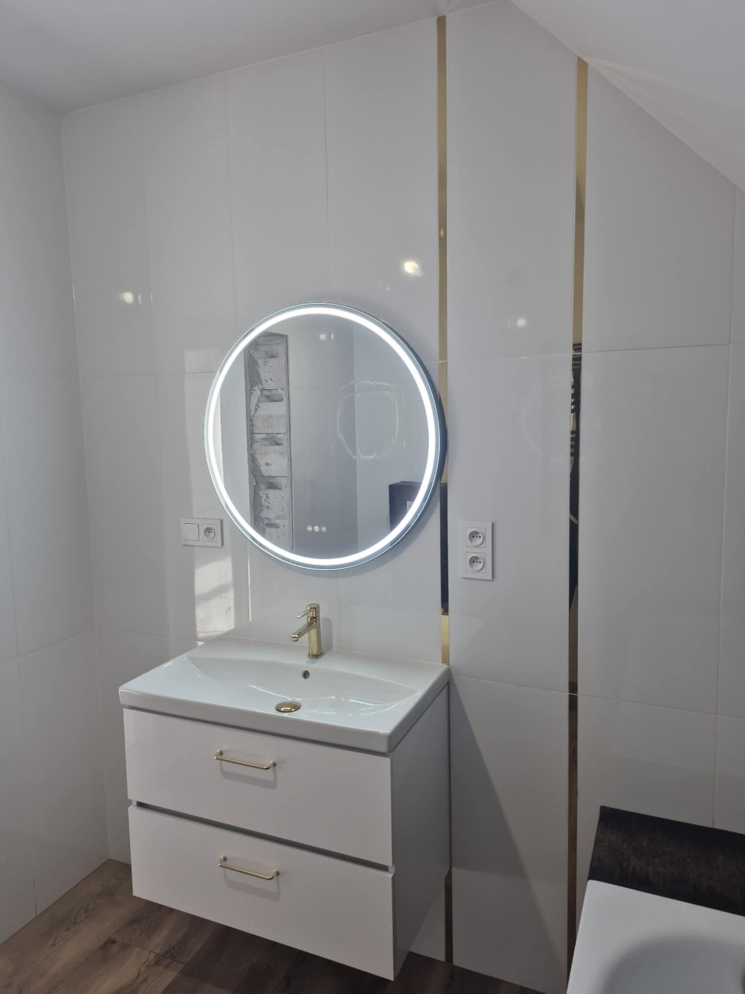 Biała łazienka z umywalką na szafce, okrągłym lustrem z podświetleniem LED oraz złotymi akcentami na białych płytkach ściennych.