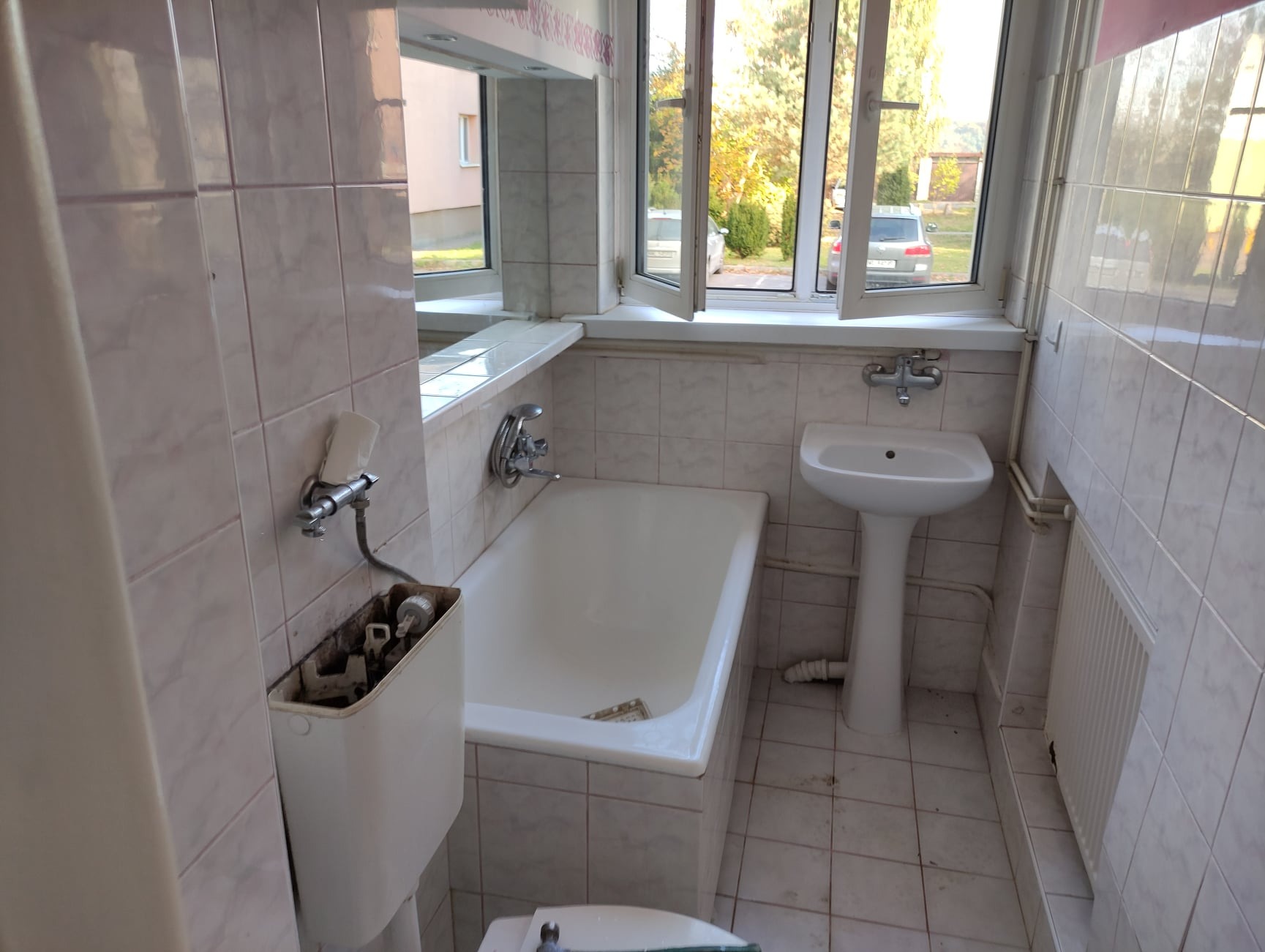 Łazienka w trakcie demontażu: widoczna wanna, umywalka na postumencie, częściowo zdemontowana spłuczka toalety, jasne płytki na ścianach i podłodze, okno z widokiem na drzewa.