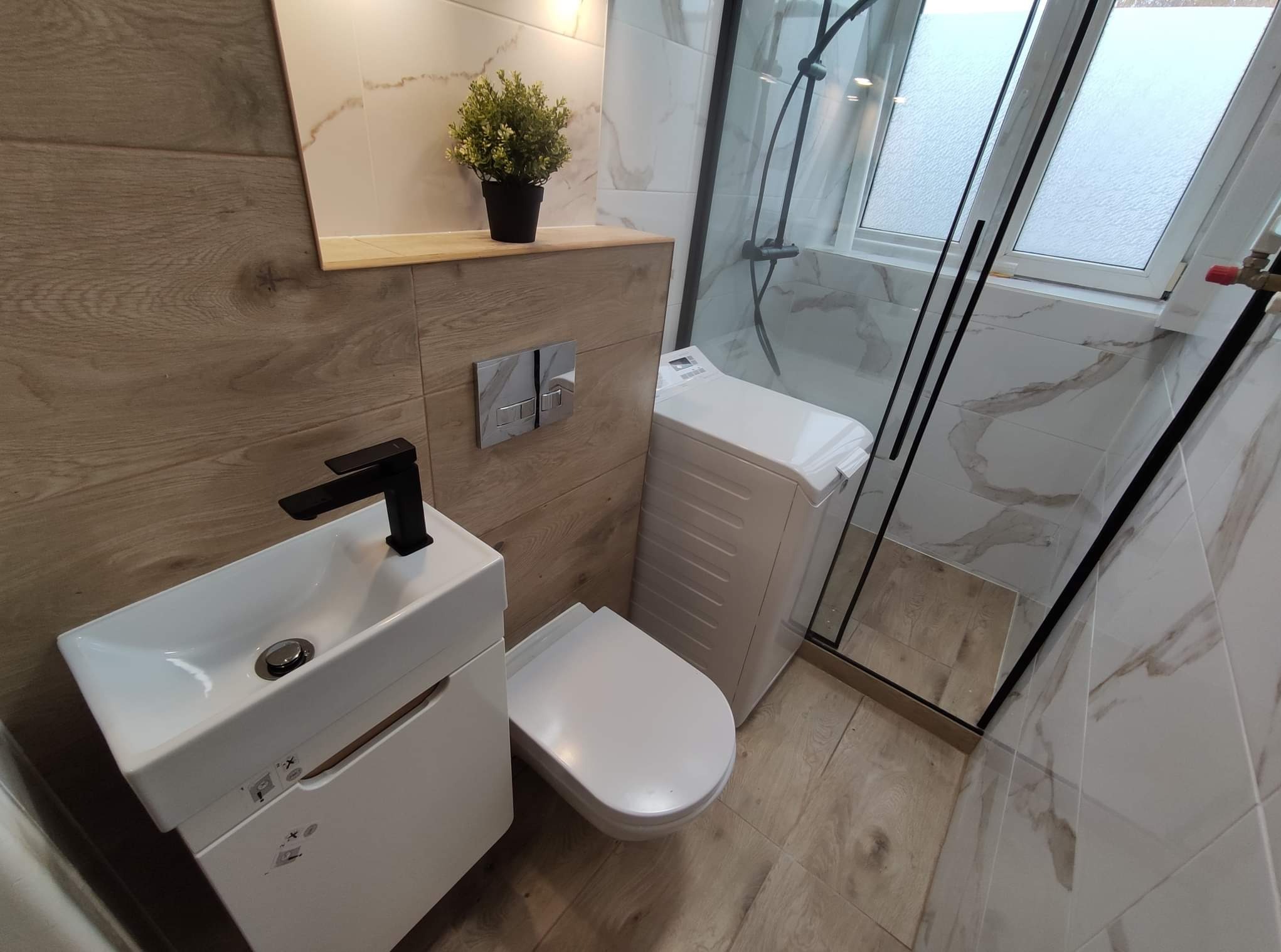 Nowoczesna łazienka z drewnopodobnymi i marmurkowymi płytkami, wisząca toaleta, umywalka z czarną baterią oraz kabina prysznicowa ze skośnym oknem dachowym.