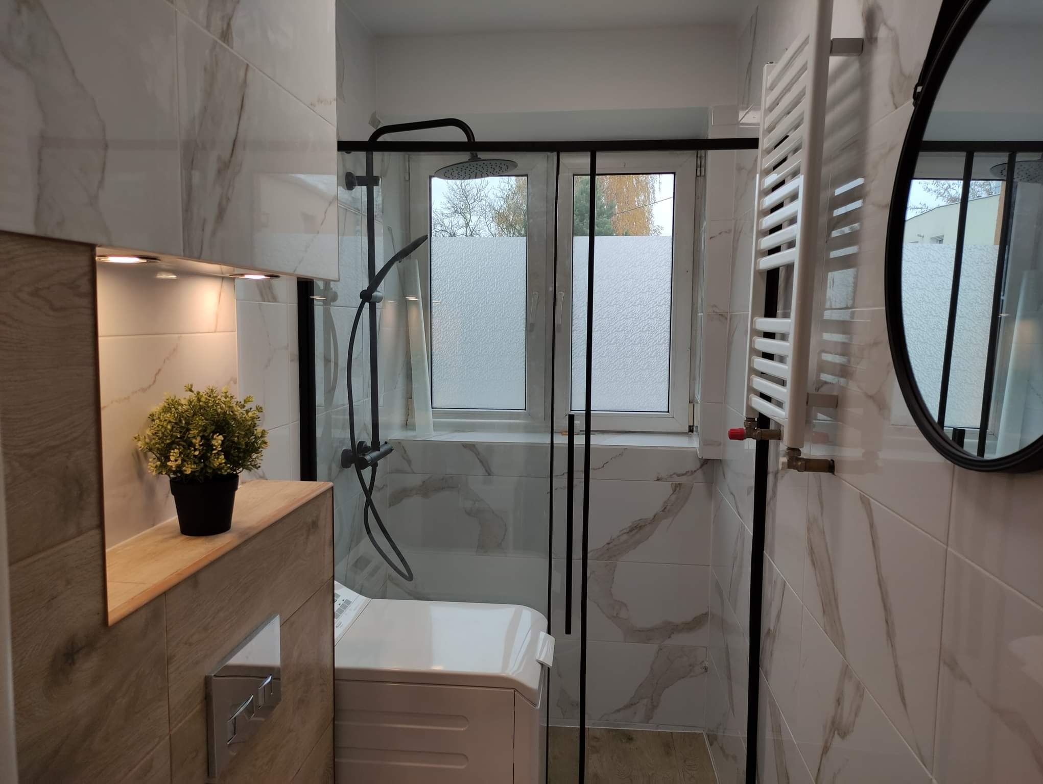 Nowoczesna łazienka z kabiną prysznicową typu walk-in, wykończona płytkami imitującymi marmur, z czarnymi elementami armatury i grzejnikiem drabinkowym; widoczna pralka i roślina doniczkowa.