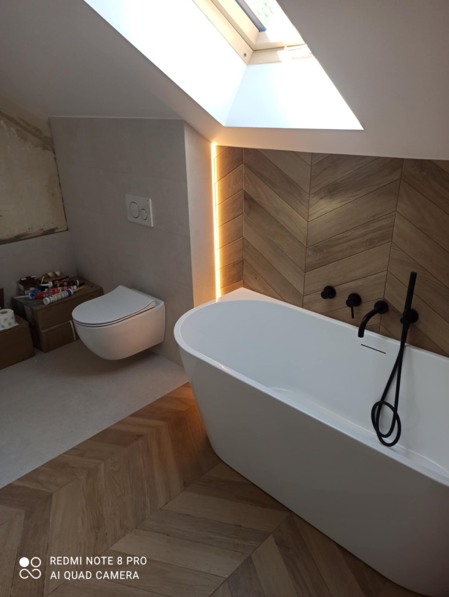 Nowoczesna łazienka na poddaszu z drewnianą podłogą ułożoną w jodełkę, białą wanną wolnostojącą z czarną armaturą, podwieszaną toaletą i oknem dachowym.