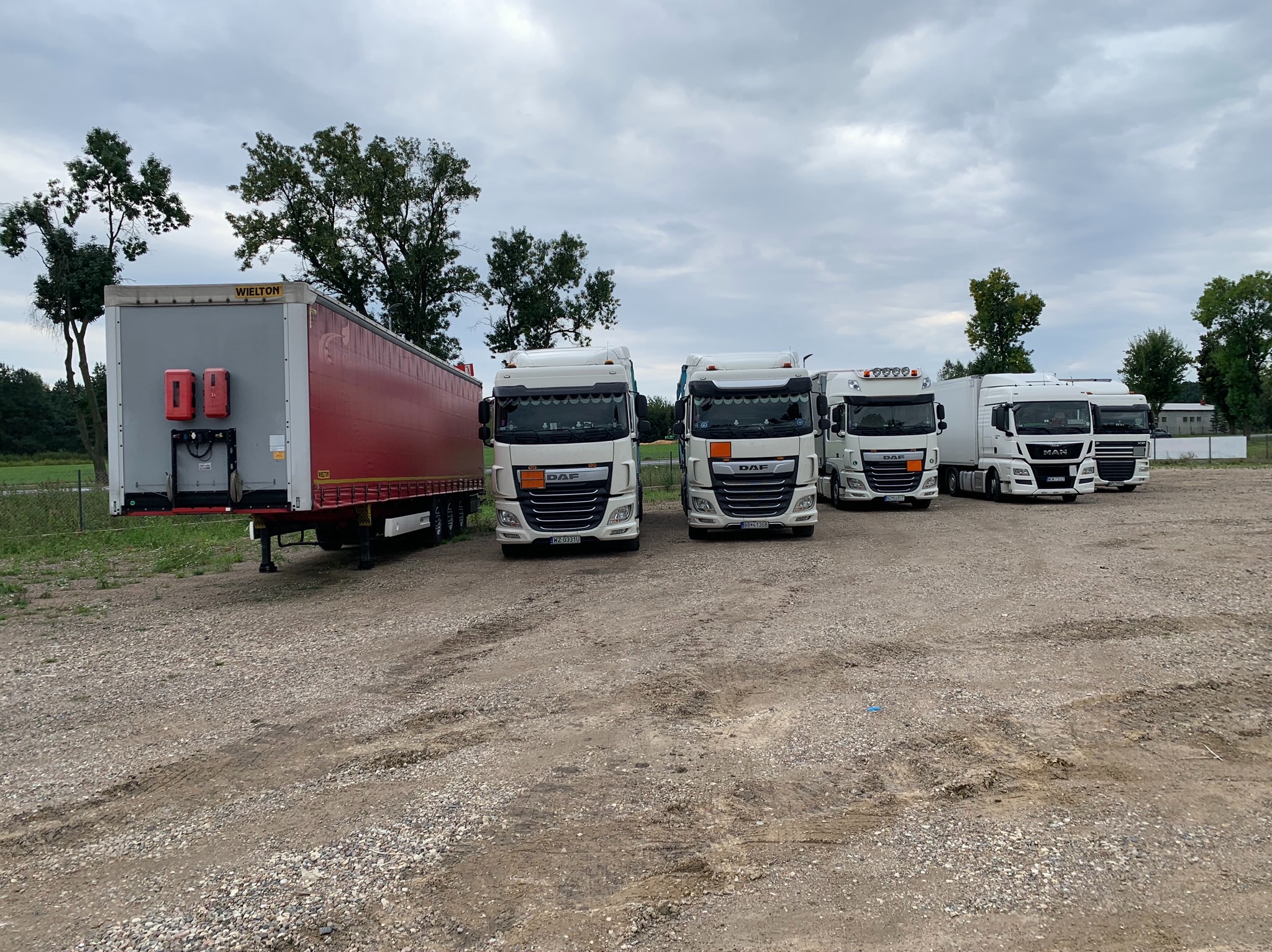 Rząd zaparkowanych białych ciężarówek DAF i MAN oraz czerwona naczepa Wielton na żwirowym placu pod pochmurnym niebem.