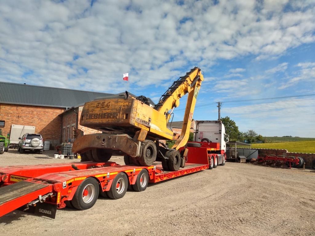 Żółta koparka Liebherr 924 na czerwonej niskopodwoziowej naczepie Faymonville, przewożona przez ciągnik Scania, na tle budynku z cegły i pola uprawnego pod błękitnym niebem.