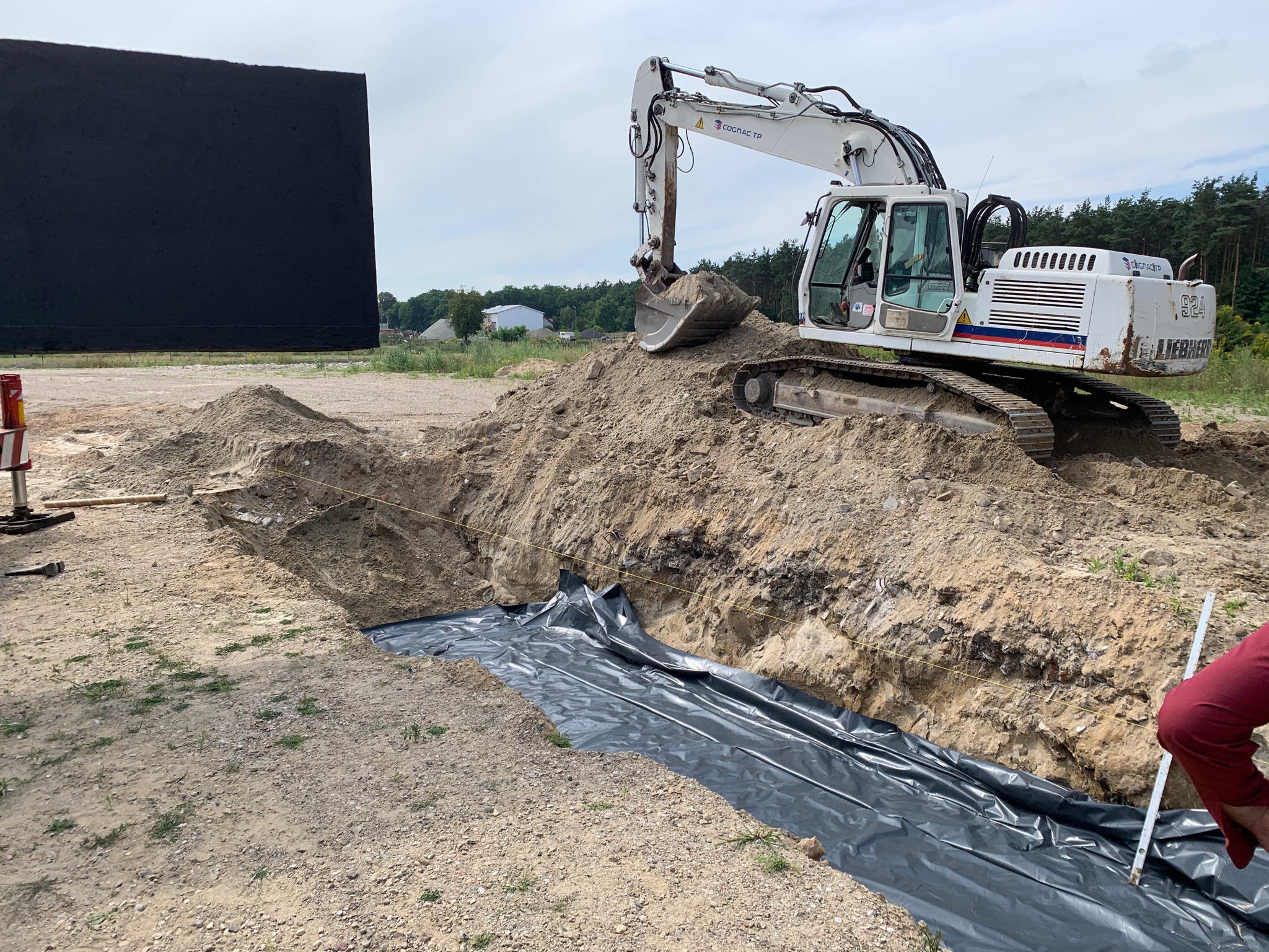 Biała koparka Liebherr na usypanym wzgórzu ziemi obok wykopu wyłożonego czarną folią, w tle drzewa i budynki.