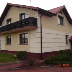 Dwukondygnacyjny dom z beżową elewacją, ciemnym balkonem z ozdobną balustradą i czerwoną dachówką, widok z poziomu chodnika z kostki.