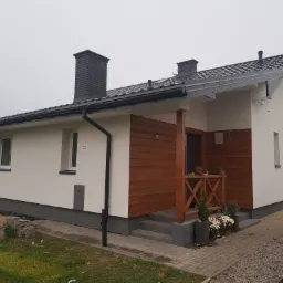 Nowoczesny dom jednorodzinny z białą elewacją, drewnianą okładziną, ciemną podmurówką i szarym dachem, widoczny z zewnątrz, z dwoma kotami siedzącymi na balustradzie werandy.