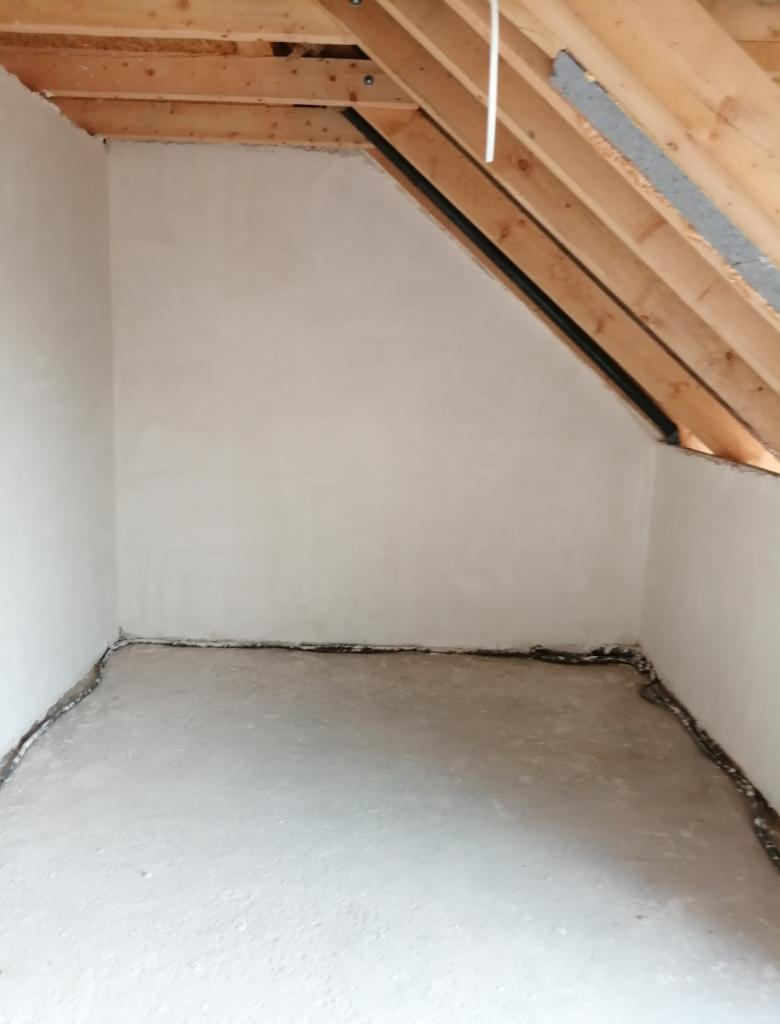 Surowe wnętrze poddasza z drewnianą konstrukcją dachu i świeżo nałożonym tynkiem na ścianach, widoczne zabezpieczenie folią przy podłodze.