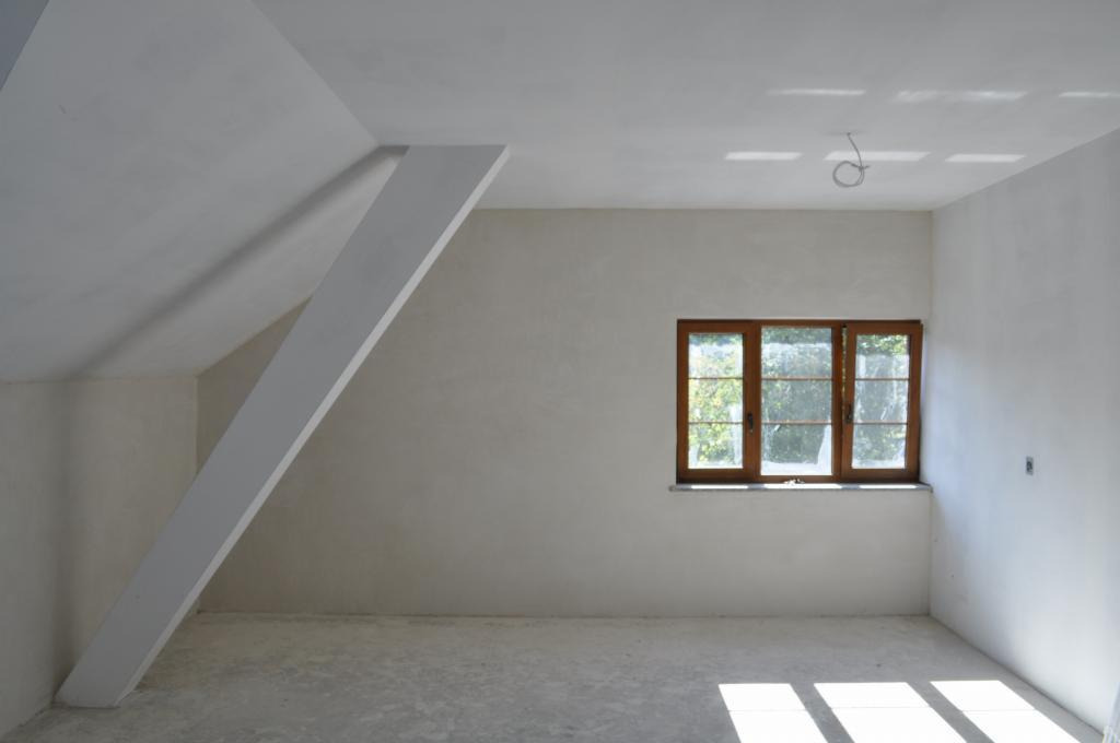 Wnętrze poddasza po tynkowaniu maszynowym, widoczny skośny sufit, drewniane okno i surowy beton na podłodze.