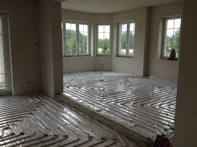 Wnętrze w stanie surowym z rozłożonymi rurami ogrzewania podłogowego, widoczne okna z widokiem na zieleń i fragmenty ścian przygotowanych pod tynkowanie.