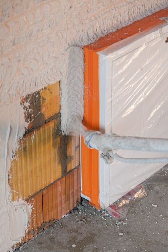 Aplikacja gładzi gipsowej na ścianę z cegły za pomocą agregatu natryskowego; okno zabezpieczone folią.