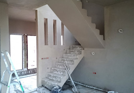 Wnętrze w trakcie wykańczania: schody i ściany pokryte świeżym, jasnym tynkiem, widoczne ślady prac, drabina oparta o ścianę, surowy stan wykończenia.