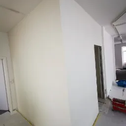 Świeżo wykończony narożnik w pomieszczeniu po pracach remontowych, zabezpieczone podłogi, widoczny fragment futryny drzwiowej i wózek malarski w tle.