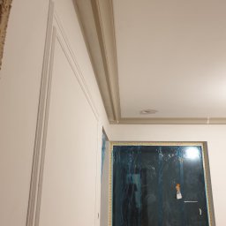 Jarosław Florkiewicz - Wykończona ściana z dekoracyjnymi listwami i gzymsem sufitowym. Okno zabezpieczone folią malarską. Widoczne detale wykończeniowe.