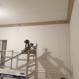 Jarosław Florkiewicz - Wykończone ściany z dekoracyjną sztukaterią i listwami, widoczne rusztowanie z narzędziami. Wnętrze w trakcie prac wykończeniowych.