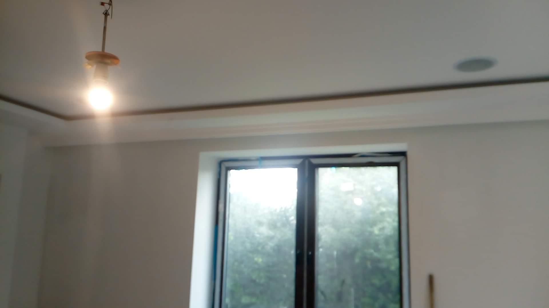 Wnętrze pokoju po remoncie, widoczne gładkie ściany pomalowane na biało, okno z widokiem na zieleń oraz lampa wisząca z żarówką.