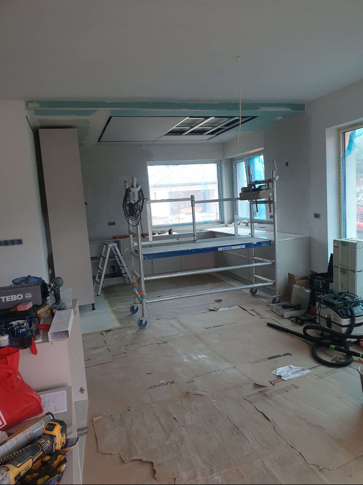 Wnętrze w trakcie montażu sufitu podwieszanego z płyt kartonowo-gipsowych. Widoczne rusztowanie, narzędzia i zabezpieczona podłoga. Częściowo zamontowane płyty.