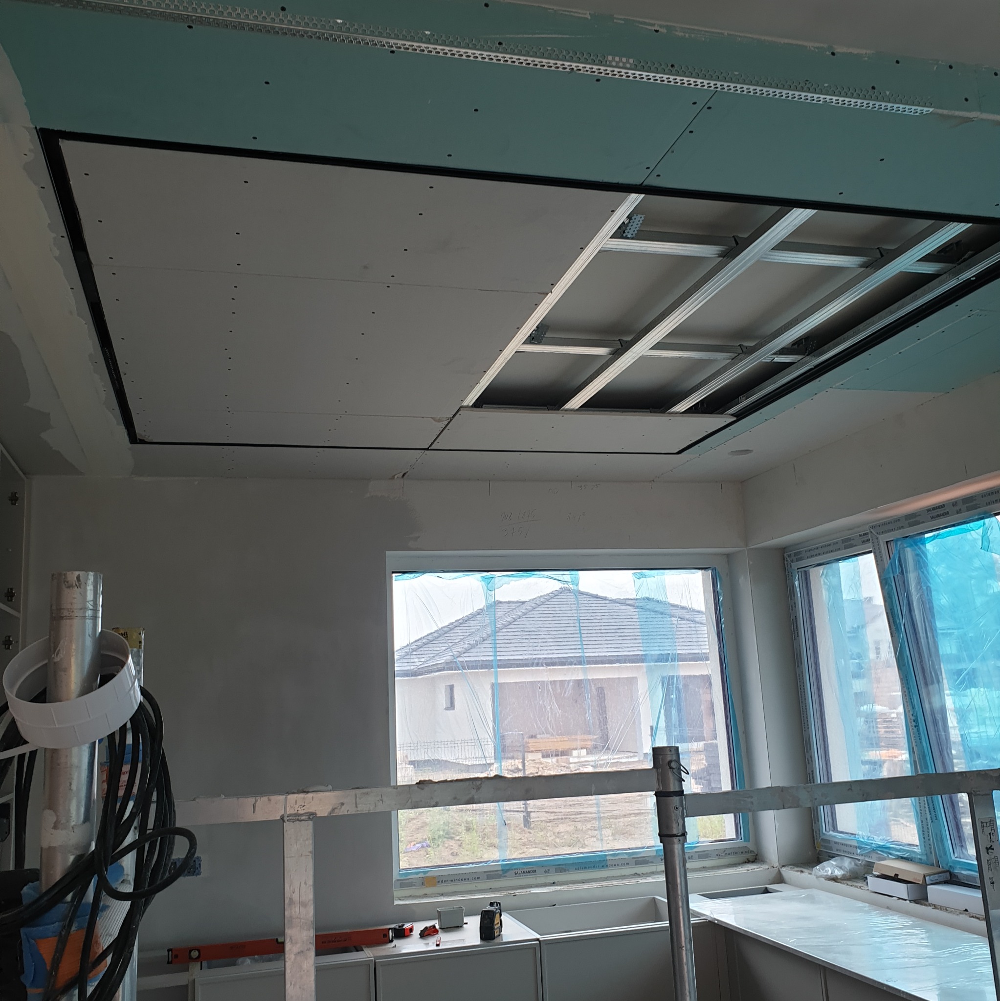Sufit podwieszany z płyt karton-gips, widoczna konstrukcja metalowa. Okno z widokiem na dom w tle. Etap prac wykończeniowych, wnętrze w stanie surowym.