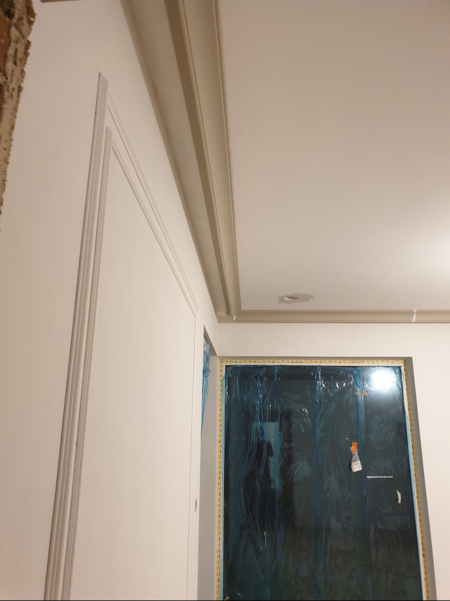 Wykończona ściana z dekoracyjnymi listwami i gzymsem sufitowym. Okno zabezpieczone folią malarską. Widoczne detale wykończeniowe.