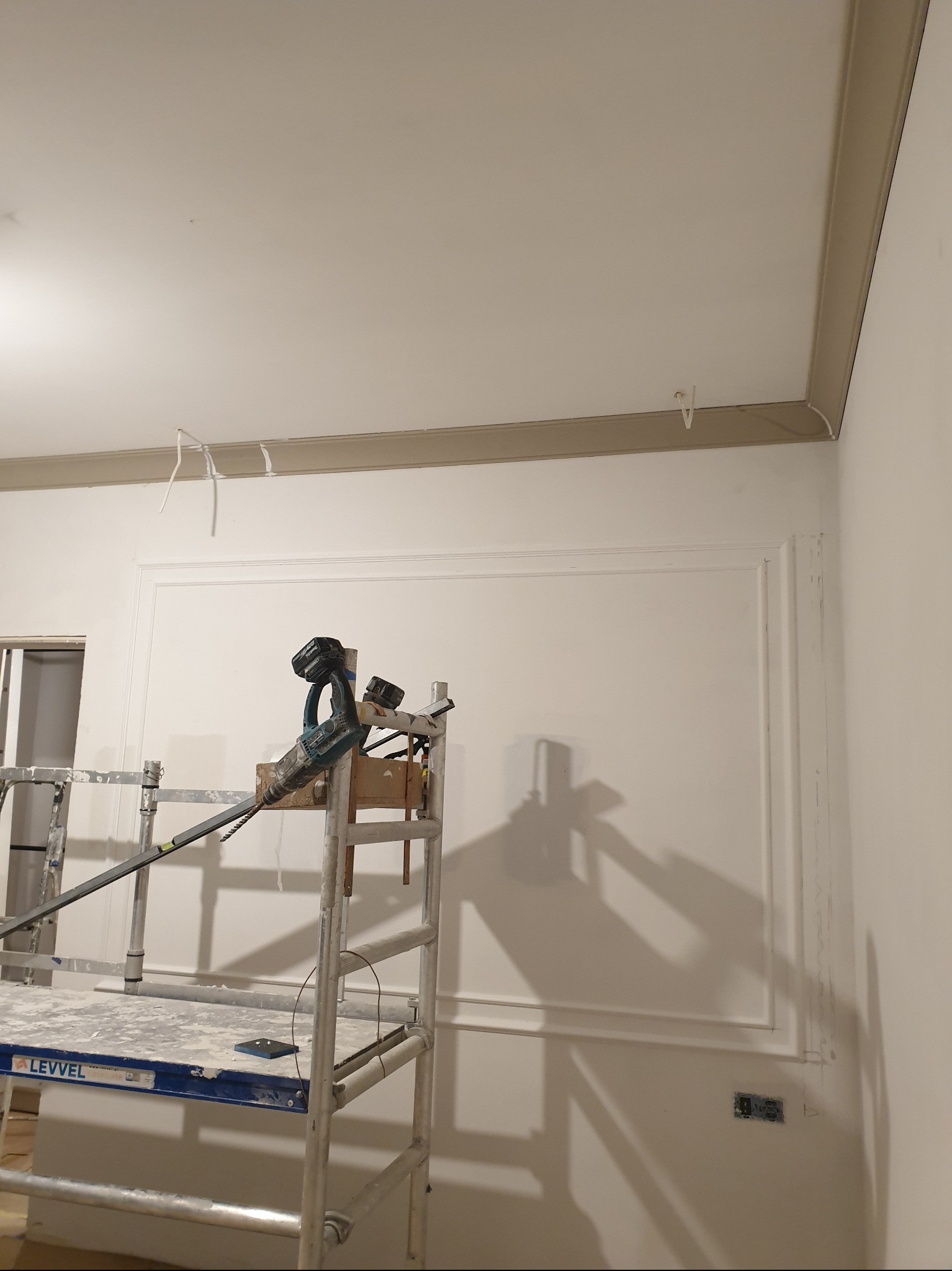 Wykończone ściany z dekoracyjną sztukaterią i listwami, widoczne rusztowanie z narzędziami. Wnętrze w trakcie prac wykończeniowych.