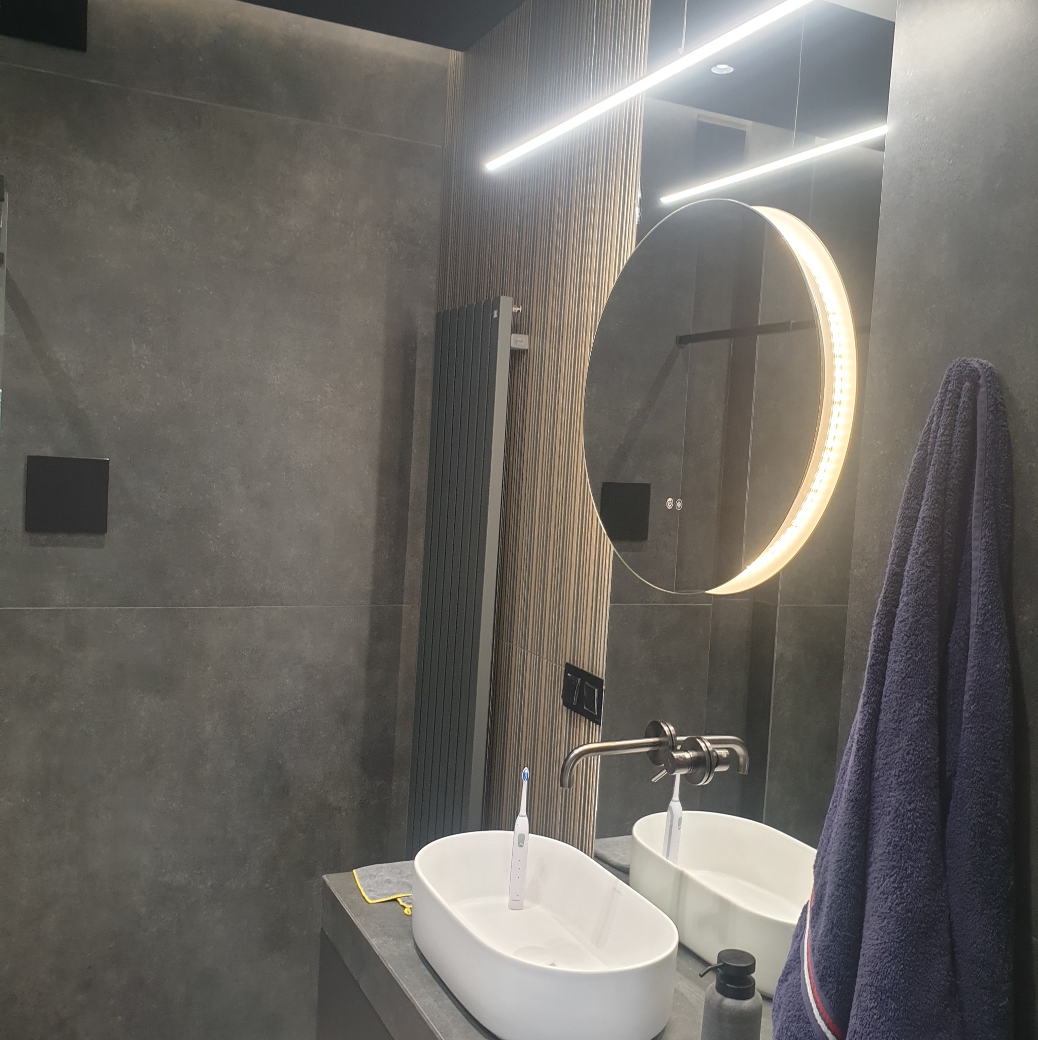 Nowoczesna łazienka w odcieniach szarości z owalnym lustrem podświetlonym LED, umywalką nablatową i grzejnikiem drabinkowym na tle ściany z płytek.