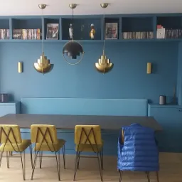 Niebieska zabudowa meblowa z półkami na książki nad stołem jadalnym, oświetlona trzema wiszącymi lampami w stylu retro, z żółtymi krzesłami i niebieską kurtką na jednym z nich.