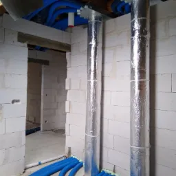 Instalacja rekuperacji w surowym budynku: widoczne dwa pionowe, zaizolowane przewody wentylacyjne z podłączonymi niebieskimi rurami wentylacyjnymi, ściany z białego betonu komórkowego.