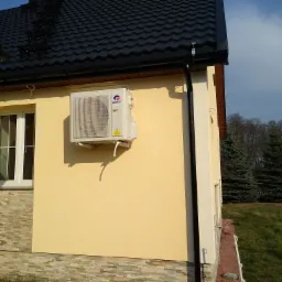 Zewnętrzna jednostka klimatyzatora marki Gree zamontowana na elewacji domu z żółtym tynkiem i kamienną podmurówką, widok z boku na tle ciemnej dachówki i zielonego trawnika