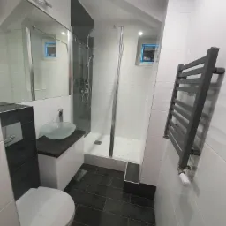 Wąska łazienka z kabiną prysznicową, szklaną umywalką na blacie, grzejnikiem drabinkowym i ciemną podłogą z płytek imitujących drewno.