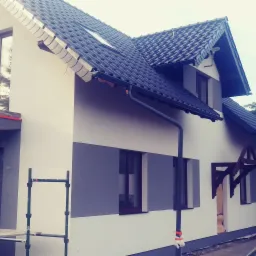 Dwukondygnacyjny dom z nową elewacją w odcieniach bieli i szarości, widoczne rusztowania i zabezpieczenia okien folią, co wskazuje na trwające prace wykończeniowe.