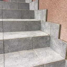 Montaż szarych, kwadratowych płytek na schodach zewnętrznych, widoczne plastikowe krzyżyki dystansowe, ściana wykończona czerwonawym tynkiem.