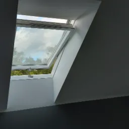 Otwarty, biały okno dachowe w szarej ścianie poddasza, widok na pochmurne niebo i drzewa.