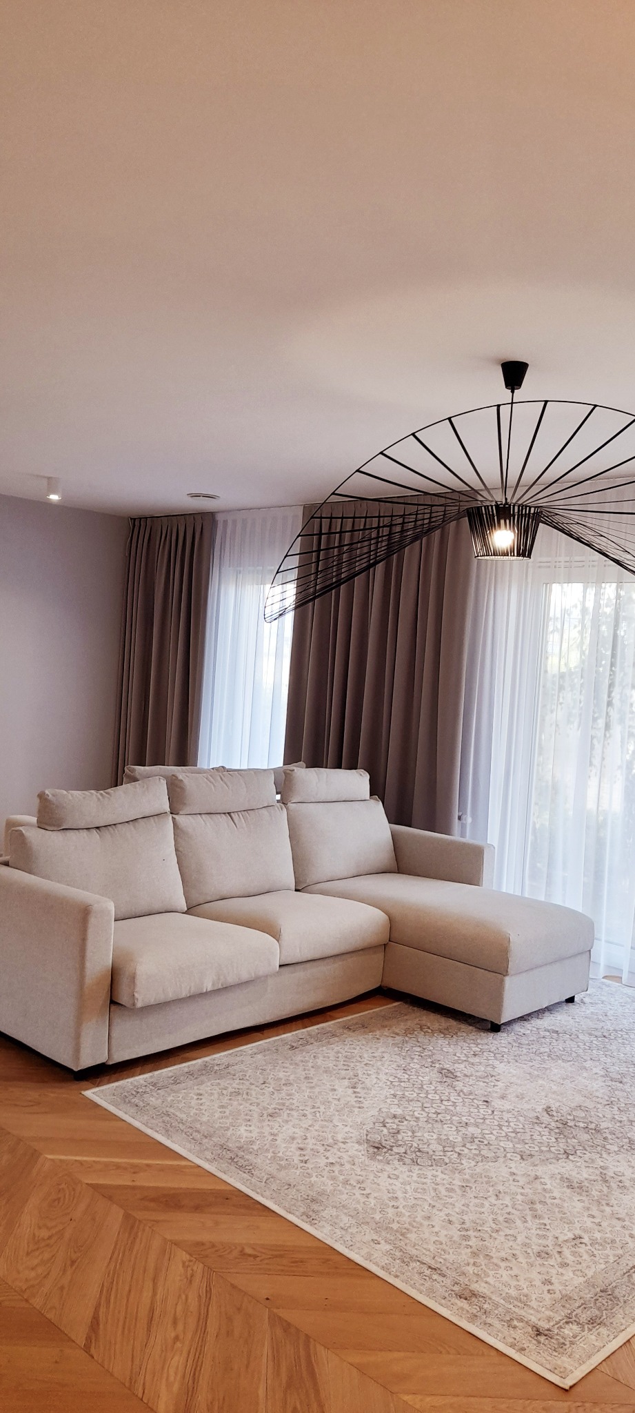 Beżowa sofa narożna z regulowanymi zagłówkami, na wzorzystym dywanie, w tle brązowe zasłony i ażurowa lampa sufitowa.