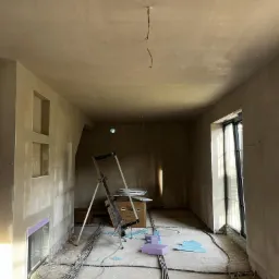 Wnętrze w trakcie prac wykończeniowych: ściany i sufit po nałożeniu szarej warstwy, widoczne kable na podłodze, drabina, okno i resztki materiałów budowlanych.