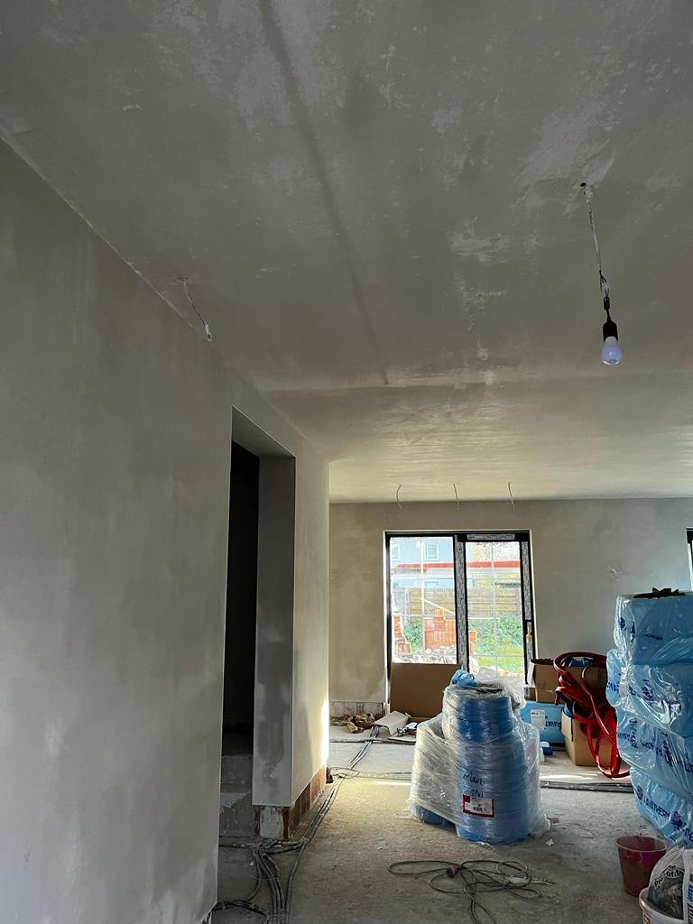 Surowe wnętrze w trakcie prac wykończeniowych: ściany pokryte szarą warstwą, widoczne przewody elektryczne, okno z widokiem na ogród, materiały budowlane zabezpieczone folią.