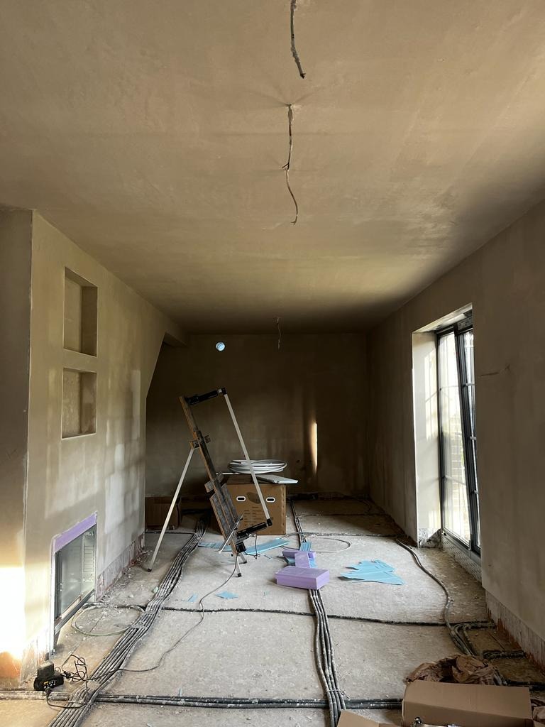 Wnętrze w trakcie prac wykończeniowych: ściany i sufit po nałożeniu szarej warstwy, widoczne kable na podłodze, drabina, okno i resztki materiałów budowlanych.